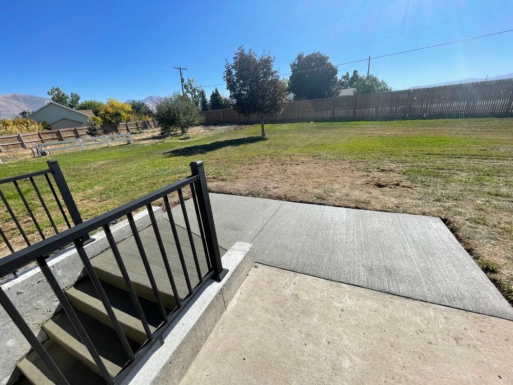 View of a grassy yard with trees and a concrete patio steps under a clear blue sky.