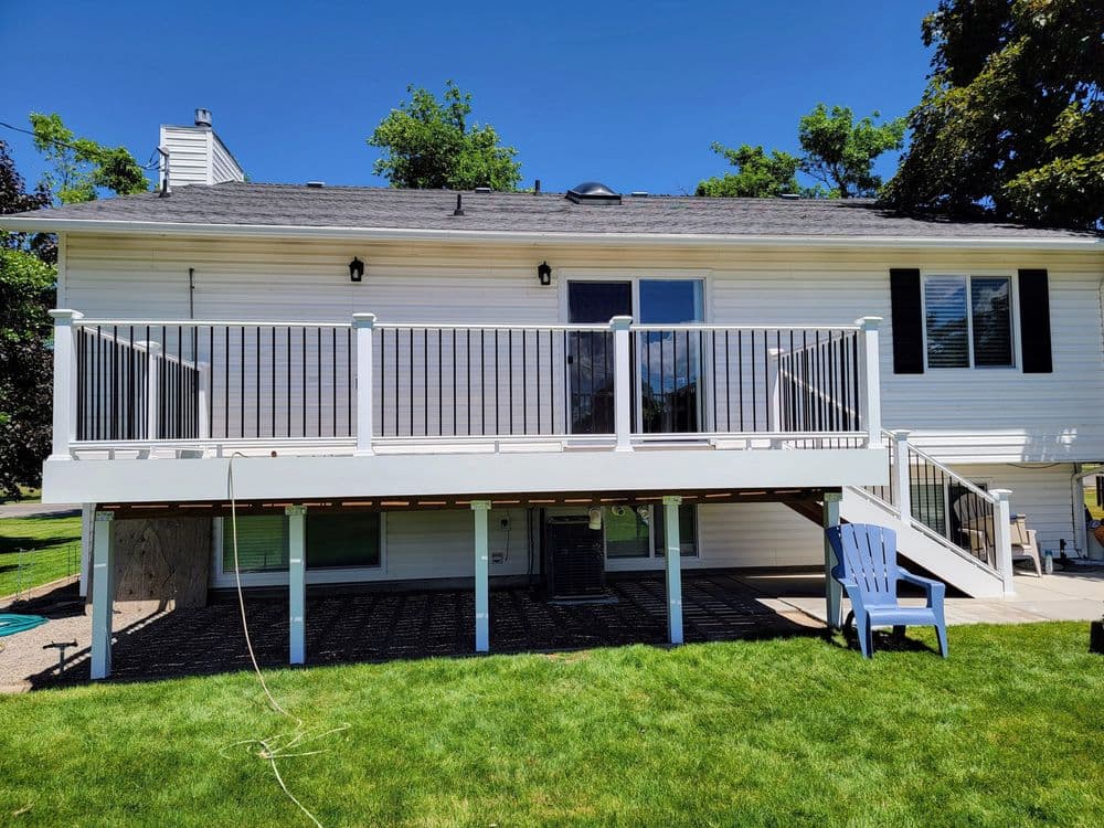 Two-story house with a white deck, green lawn, and blue chair under clear sky.